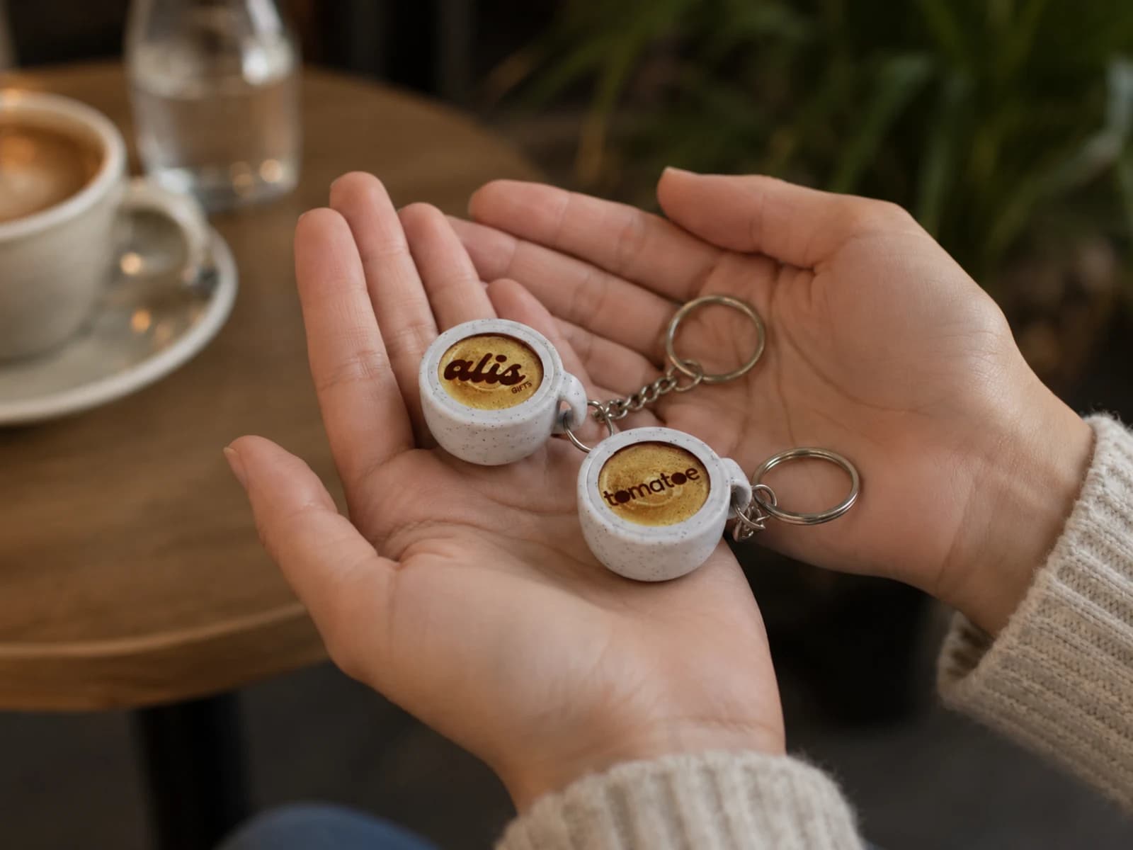 Dos llaveros con forma de taza de café en la palma de una mano, uno con el logo de alis GIFTS y otro con el de tomatoe impresos sobre el café.